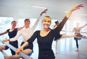 Dancers in a studio perform controlled leg stretches beside ballet barres before class.